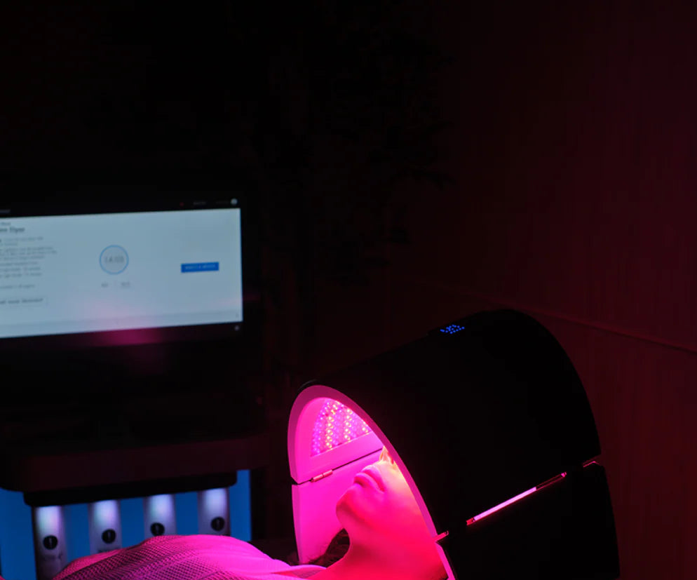Person lying on a massage table with pink light therapy device on feet in a dimly lit room.